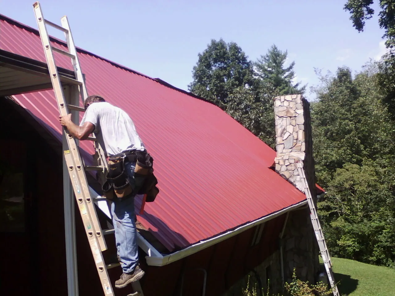 Roofer on a ladder beside a red metal roof and stone chimney — Soffit & Fascia Repair by Roofing Brothers in Guerneville